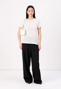 Bílé perforované pletené tričko s krátkými rukávy, v kombinaci s volnými černými kalhotami. Modelka nese malou bílou kabelku a má na sobě černé slip-on boty.