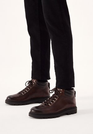 Botas de senderismo de cuero marrón oscuro con acabado texturizado, ojales de metal y suelas de goma negra, combinadas con pantalones negros.