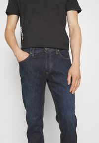 Ciemnoniebieskie dżinsy o prostym kroju, z pomarańczowym przeszyciem, przednimi kieszeniami i zapięciem na guzik. Połączone z czarnym t-shirtem.