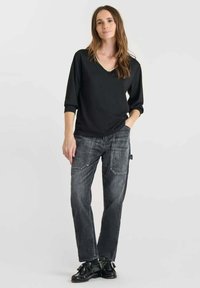 Donna con lunghi capelli castani, che indossa una maglietta nera a V, jeans grigi con toppe e scarpe di pelle nera con lacci, in piedi davanti a uno sfondo semplice.