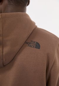 Bruine hoodie met een zwart logo van "The North Face" op de voorkant. De stof lijkt zacht en heeft een gestructureerde, comfortabele pasvorm.