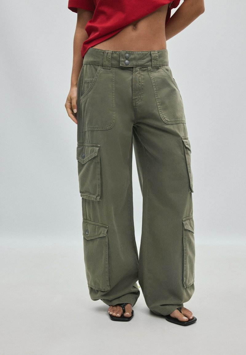 Personne portant un pantalon cargo ample vert olive avec plusieurs poches à boutons et des sandales noires à bout carré sur un fond uni.