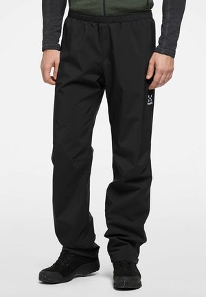 Zwarte waterdichte outdoorbroek met elastische tailleband, gedragen met zwarte schoenen en een groen-grijs longsleeve shirt.