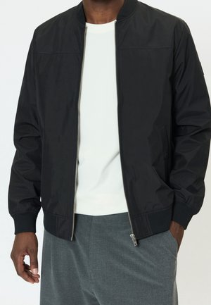 Homme portant une veste bomber noire zippée sur une chemise blanche et un pantalon gris, une main dans la poche et l'autre détendue sur le côté.