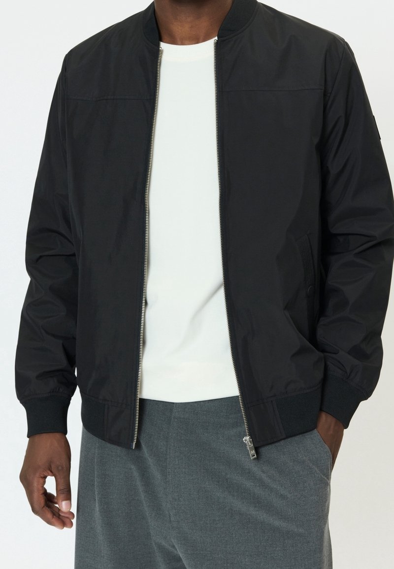 Homme portant une veste bomber noire zippée sur une chemise blanche et un pantalon gris, une main dans la poche et l'autre détendue sur le côté.