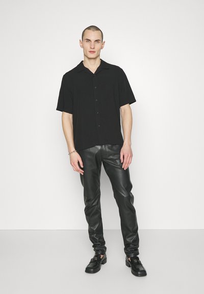 Schwarzes kurzärmeliges Hemd mit Knopfleiste und kragen, kombiniert mit eng anliegenden schwarzen Lederhosen und schwarzen Schuhen. Glatte Textur, minimalistisches Design.