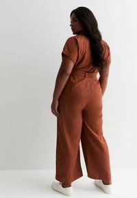 Combinaison couleur rouille avec une coupe à jambes larges, des manches courtes et une taille ceinturée. Le modèle porte des baskets blanches. Tissu lisse et léger.