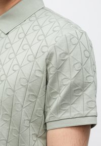 Ανοιχτοπράσινο πόλο με υφασμένο, ανάγλυφο σχέδιο που περιλαμβάνει ρόμβους και καμπυλωτά διακοσμητικά στοιχεία. Κοντά μανίκια με ριμπ μανσέτες.