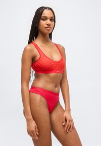 Γυναίκα με μακριά πλεξούδες που φοράει κόκκινο αθλητικό σουτιέν Adidas και αντίστοιχα μαγιό, στέκεται μπροστά σε απλό ανοιχτό γκρι φόντο.