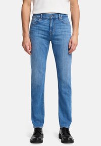 Niebieskie dżinsy w stylu prostym, wykonane z denimu, z wyblakłym wykończeniem, pięcioma kieszeniami i zapięciem na guzik. Noszone z czarnymi butami.