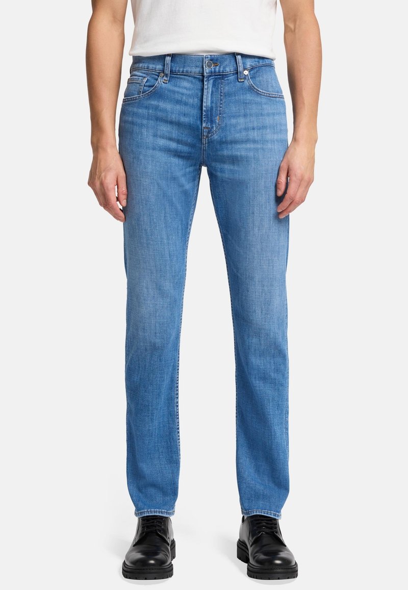 Niebieskie dżinsy w stylu prostym, wykonane z denimu, z wyblakłym wykończeniem, pięcioma kieszeniami i zapięciem na guzik. Noszone z czarnymi butami.