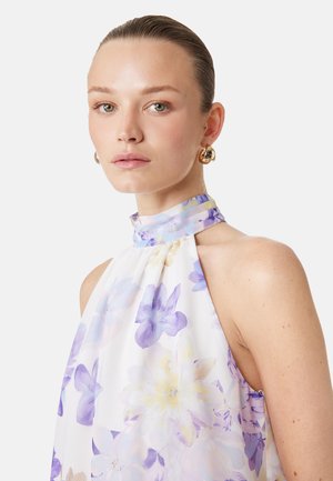 Jeune femme aux cheveux plaqués en arrière, portant des boucles d’oreilles créoles dorées et une robe blanche sans manches à imprimé floral violet et jaune.