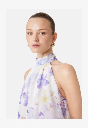 Jeune femme aux cheveux plaqués en arrière, portant des boucles d’oreilles créoles dorées et une robe blanche sans manches à imprimé floral violet et jaune.