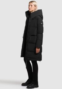 Parka nera imbottita con collare oversize, vita regolabile con cordoncino e tasche frontali. Chiusura con zip e patch con logo sulla manica.