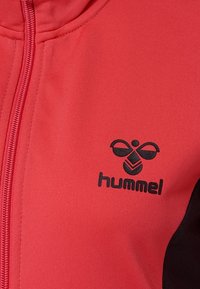 Veste zippée rouge avec des accents noirs. Présente une texture de tissu lisse et un logo "hummel" noir avec un design unique sur la poitrine.