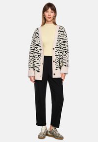 Leichte Strickjacke in Creme mit schwarzen Tigerstreifen, ausgestattet mit einem pinkfarbenen, gerippten Rand, vier Knöpfen und Seitentaschen. Kombiniert mit schwarzen Hosen und gemusterten Schuhen.