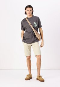 Camisa de algodón gris con gráficos en verde claro, pantalones cortos beige y zapatillas marrones. Modelo llevando un bolso cruzado marrón, de pie frente a un fondo liso.
