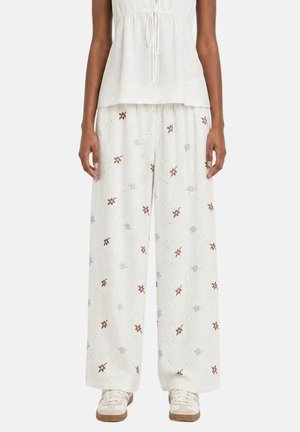 Mannequin portant un pantalon blanc ample avec de petits motifs floraux marron et bleu clair, associé à un haut blanc sans manches et des baskets blanches.