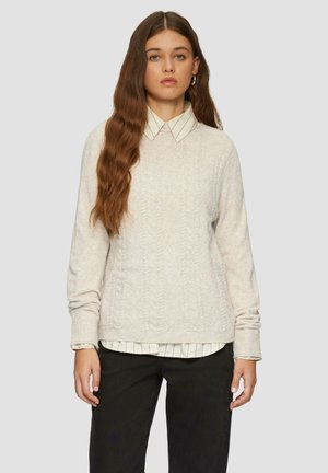 Pull en laine beige clair à mailles torsadées avec un col rond et des poignets côtelés, superposé à une chemise blanche à rayures noires, associé à un pantalon foncé.