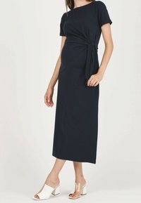 Marinblå gravidklänning med korta ärmar, knuten midjedetalj och en svävande silhuett, parat med tvårands blockklacksandaler.