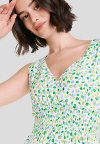 El vestido de estampado floral presenta manchas verdes y azules sobre un fondo blanco, con un escote en V, diseño sin mangas y detalle de cintura fruncida.