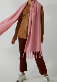 Personne portant une chemise en cuir marron, un pantalon rouge foncé, des baskets blanches et une longue écharpe rose à franges drapée sur les épaules, sur fond uni.