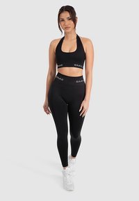 Czarny sportowy biustonosz i legginsy z wysokim stanem z logo, wykonane z elastycznego materiału. Modelka pozuje w białych sneakersach.