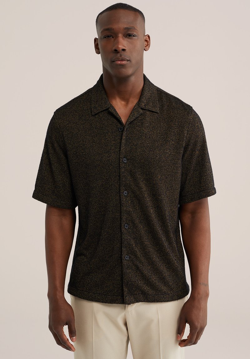 Homme portant une chemise à manches courtes marron foncé texturée avec boutons, et un pantalon beige, debout devant un fond clair uni.