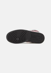 Semelle en caoutchouc noire d'une basket Nike avec des motifs de bande de roulement circulaires et rectangulaires, visible par le dessous sur un fond clair uni.