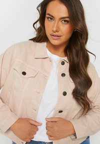 Vaaleanpunainen farkkutakki, jossa on perinteinen kaulus, etutaskut ja mustat napit. Pehmeä rakenne ja sovitettu malli luovat ryhdikkään siluetin.