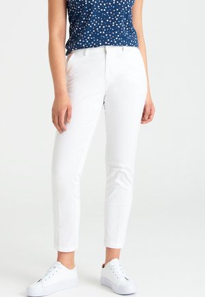 Vrouw draagt strakzittende witte broek, witte sneakers en een marineblauw mouwloos topje met een klein bloemmotief.