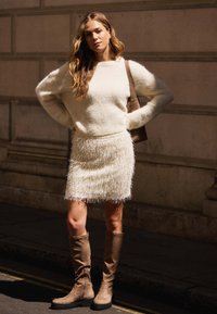 Kremowy sweter i strukturalna biała spódnica w frędzle w połączeniu z brązowymi zamszowymi botkami do kolan, z zamkiem błyskawicznym po boku i mocną podeszwą.