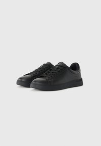 Black sneaker made of leather with a round toe, lace-up design, textured upper, and a flat rubber sole. Minimalist style with no visible logos.