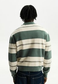 Person med kort, snurrat hår som bär en grön, vit och grårandig tröja och mörkblå jeans, vänd bort från kameran mot en enkel vit bakgrund.