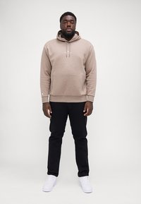 Felpa beige con tasca a marsupio, cappuccio regolabile con cordino e logo bianco. Abbinata a pantaloni neri e sneaker bianche.