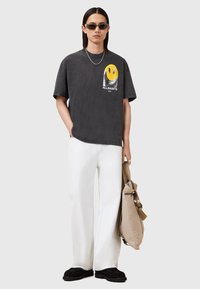 Grafisk grå T-shirt med et gult smilende ansigt logo, parret med hvide vide bukser og sorte teksturerede sko, mens de holder en beige taske.