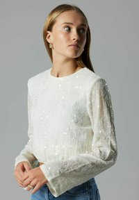 Hvid, tekstureret langærmet skjorte med rund halsudskæring, prydet med spredte perler og en let samlet kant. Kombineret med blå jeans.