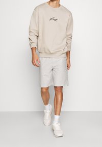 Beige sweatshirt med sort logo, lysegrå shorts og hvide sneakers. Sweatshirten har rund halsudskæring og ribbede manchetter.