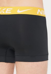 Crne bokserice s žutim elastičnim pojasom na kojem se nalaze bijeli "Nike" brendirani natpisi, glatka tkanina, uski kroj i detalji šivanja.
