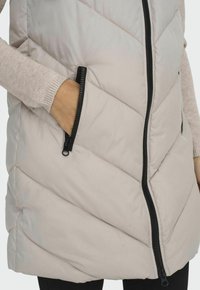 Gilet piumino leggero beige con trapuntatura a zigzag, zip frontale completa e tasche con zip nere. Texture morbida e design minimalista.