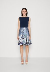 Navy bodice dress with a flared blue floral print skirt. Wide neckline, sleeveless design, and knee-length, paired with white strappy heels.