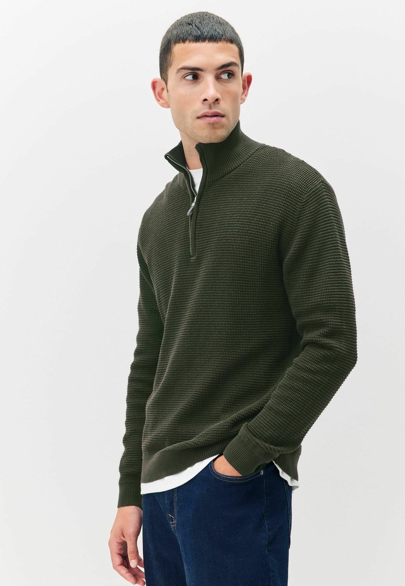 Mann trägt einen dunkelgrünen strukturierten Halbzip-Pullover, ein weißes Unterhemd und eine blaue Jeans, schaut nach rechts mit einer Hand in der Tasche.