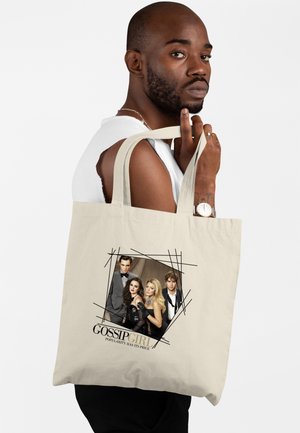Ein Mann hält eine beige Tragetasche mit vier formell gekleideten Personen, darauf steht „Gossip Girl“ und der Slogan „Popularity Has Its Price.“