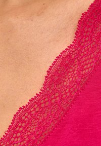 Różowy strój z głębokim dekoltem w kształcie litery V ozdobiony misterną koronkową lamówką. Materiał jest gładki i dopasowany, z teksturowanym koronkowym akcentem wzdłuż linii szyi.