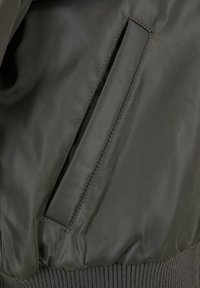 Dunkelgrüne Bomberjacke mit matter Oberfläche, gerippten Bündchen und einer Seitentasche mit Klappenverschluss. Glatte Textur und minimalistisches Design.