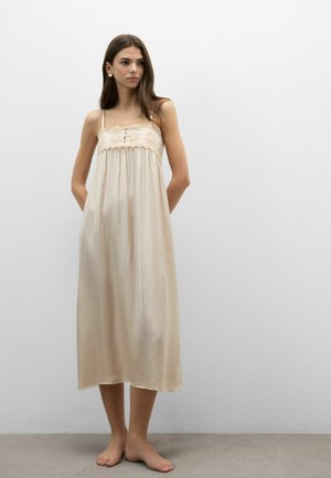 Ung kvinde med langt mørkt hår står barfodet iført en beige midi kjole med hæklet detalje ved brystet mod en almindelig hvid væg.