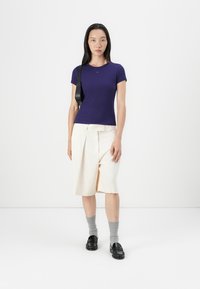 Violet trumpas rankovėmis viršus su kreminėmis klostuotomis šortais. Modelis dėvi pilkas kojines ir juodas loafer tipo batus, o taip pat turi juodą petinę rankinę.