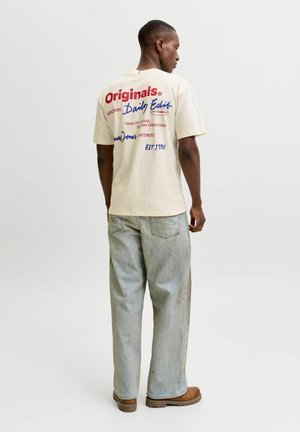 Homme debout de profil portant un t-shirt graphique beige, un jean ample clair et des bottes marron sur un fond uni.