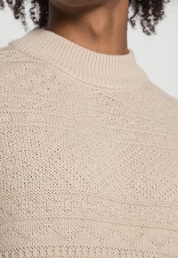 Ljusbeige stickad tröja med hög, ribbad krage. Har texturerade mönster och en slät yta som visar på intrikata stygn. 