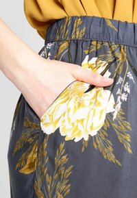 Main dans la poche d'une jupe gris foncé à imprimé floral jaune et blanc, associée à un haut jaune moutarde.